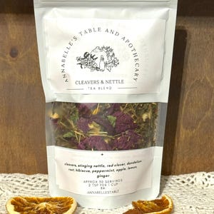 May include: A white pouch of Cleavers & Nettle tea blend with a clear window showing dried herbs and flowers. The label includes the text "ANNABELLE'S TABLE AND APOTHECARY" and a list of ingredients. Two dried orange slices are in the foreground.