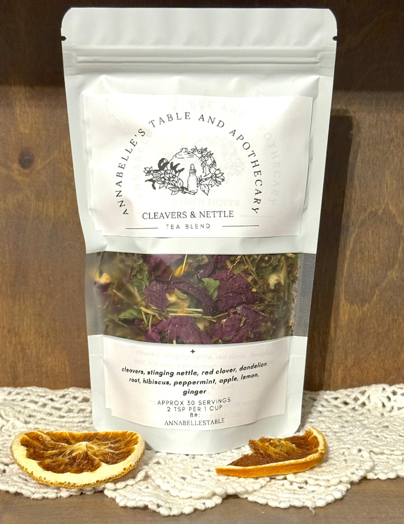 May include: A white pouch of Cleavers & Nettle tea blend with a clear window showing dried herbs and flowers. The label includes the text "ANNABELLE'S TABLE AND APOTHECARY" and a list of ingredients. Two dried orange slices are in the foreground.