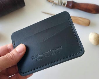May include: A black leather card holder with visible stitching along the edges. The front features the embossed text Birchmount Leather Handcrafted in Toronto. The card holder has a V-shaped pocket opening and a smooth, matte finish surface.