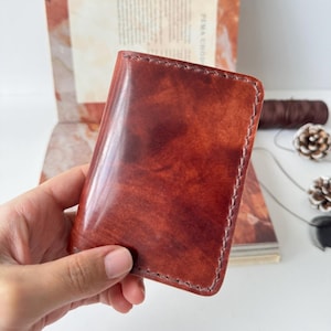 May include: A handcrafted, reddish-brown leather wallet with visible stitching around the edges. The wallet has a glossy finish and appears to be new. The background includes a book and crafting supplies, suggesting a handmade product.