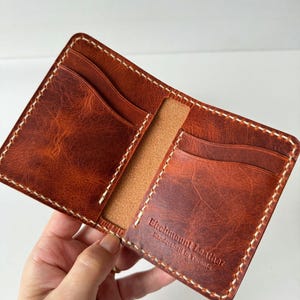 May include: Open, brown leather wallet with multiple card slots and a central pocket. The wallet is stitched with white thread and features the embossed text "Elchmount Leather handcrafted in Toronto."