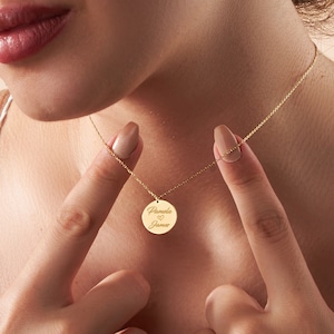 May include: A gold necklace featuring a circular pendant engraved with the names "Pamela" and "James", and a heart symbol. The necklace is held by a person. The chain is delicate.