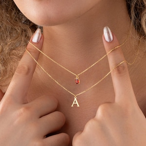 May include: Two delicate gold necklaces. The top necklace features a small, rectangular red gemstone pendant. The bottom necklace has a gold letter "A" pendant. The necklaces are layered and worn close to the neck.