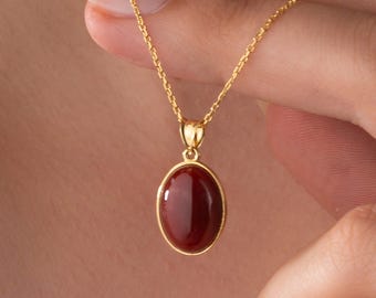 May include: A gold-colored necklace with a pendant featuring a polished, oval-shaped, reddish-brown gemstone. The pendant is set in a gold frame and hangs from a delicate gold chain. The necklace is displayed against a neutral background.