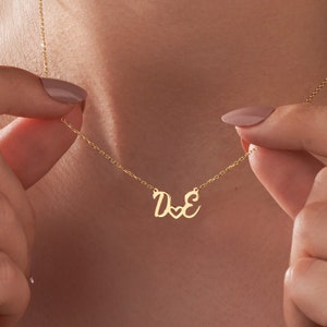 May include: Gold necklace with a cursive 'D' and 'E' connected by a heart charm.