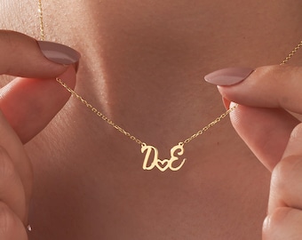 May include: Gold necklace with a cursive 'D' and 'E' connected by a heart charm.