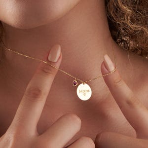 May include: Gold necklace with a round pendant that says "Mom" with a small heart charm. The charm has a pink gemstone.