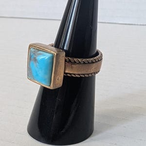 Pode incluir: Um anel em tom bronze com uma pedra quadrada azul clara. O anel é exibido em um suporte preto em forma de cone. A banda tem um design texturizado semelhante a uma corda.
