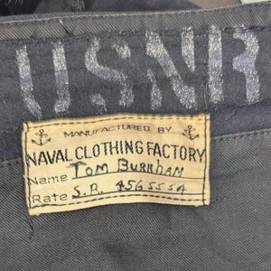May include: Black trousers featuring a Naval Clothing Factory label with the text USNR, Name Tom Burnham, and Rate S.R. 4565554. An orange tag with the number 484 is attached to the pocket flap. The fabric shows visible wear and small holes.