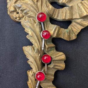 May include: Four red cabochon earrings with silver posts are displayed against a gold-toned, ornate, decorative background. The earrings have a round, shiny, red glass-like appearance. The background has a detailed, vintage, floral design.