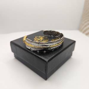 Peut inclure: Un bracelet manchette en argent et or en forme de plume dans une boîte cadeau noire. Le bracelet est en métal et présente un motif de plume délicat.
