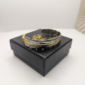 Peut inclure: Un bracelet manchette en argent et or en forme de plume dans une boîte cadeau noire. Le bracelet est en métal et présente un motif de plume délicat.