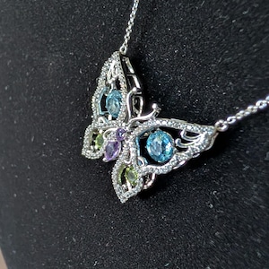 Può includere: Una collana con pendente a farfalla argentata con una catena delicata. La farfalla presenta un design a filigrana ed è adornata con pietre preziose blu, verdi e viola. La collana è esposta su uno sfondo nero.