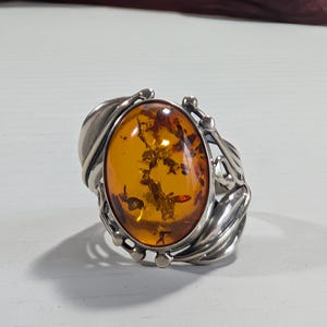 Peut inclure: Bague en argent ornée d'une grande pierre d'ambre ovale. L'ambre est d'une couleur dorée chaude, sertie dans une monture en argent détaillée avec des motifs en forme de feuilles. La bague est présentée sur une surface blanche.
