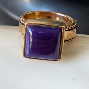 Pode incluir: Um anel dourado com uma pedra quadrada roxa escura. O anel tem uma banda texturizada e uma moldura quadrada ao redor da pedra. O anel está sobre uma superfície branca.
