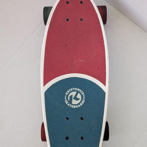 May include: A red, white, and blue Kryptonics skateboard. The top of the board is divided into red and blue sections with a white border. The Kryptonics logo is in the blue section. The skateboard has black wheels.