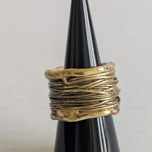 May include: A gold-toned ring with a textured, wrapped design. The ring is displayed on a black cone-shaped stand. The ring's design features multiple layers of thin, wire-like elements, creating a unique, artistic look.
