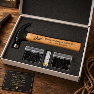May include: A personalized hammer gift set for Dad, presented in a white box with black foam. The hammer has a wooden handle engraved with "Dad" and a message. Includes nails and a tape measure. A card reads "Built with Love."