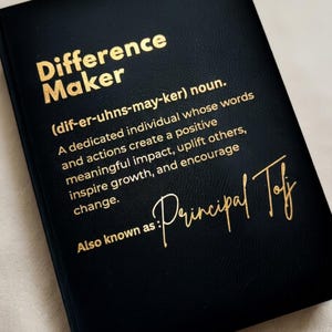 May include: A black journal with gold lettering that reads "Difference Maker." The definition of "Difference Maker" is printed below, followed by "Also known as: Principal Toff" in gold script. The journal has a textured cover.