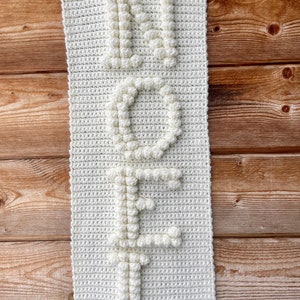 May include: White crocheted wall hanging with the word "NOEL" spelled out in raised letters.