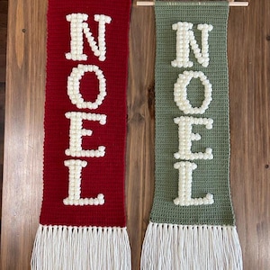 May include: Two crocheted wall hangings with the word "NOEL" spelled out in white yarn on a red and green background. Each hanging has a white fringe at the bottom.