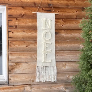 May include: White crocheted wall hanging with the word "NOEL" spelled out in white yarn pom-poms. The hanging has a fringe at the bottom and is hanging on a wooden wall.