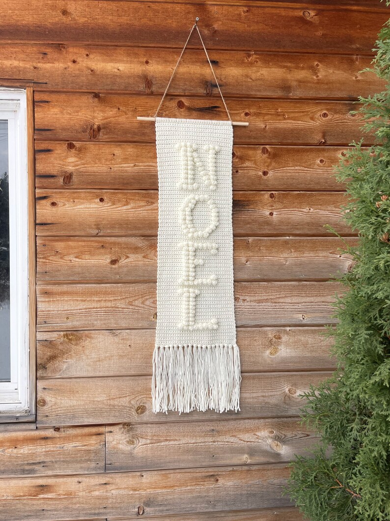 May include: White crocheted wall hanging with the word "NOEL" spelled out in white yarn pom-poms. The hanging has a fringe at the bottom and is hanging on a wooden wall.