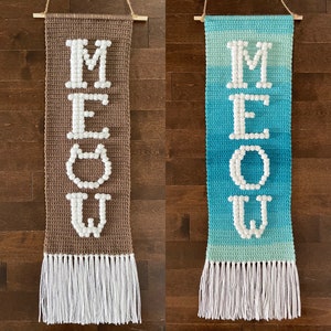 Puede incluir: Dos tapices de pared de crochet con pompones blancos que deletrean la palabra "MEOW". Uno es marrón y el otro es azul con un degradado verde.