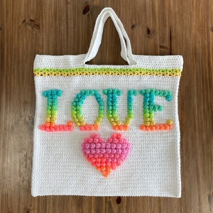 Puede incluir: Bolsa de mano de crochet blanca con un diseño de "LOVE" arcoíris y un corazón rosa.