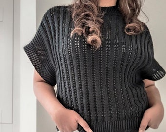 May include: Black knit top with short sleeves and a ribbed hem. The top has a crew neck and a vertical open-weave design. The model is wearing white pants and has their hands in their pockets.