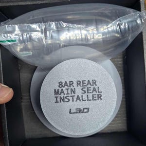 May include: An open box containing an automotive part. The main focus is a silver-colored circular disc with the text "8 BAR REAR MAIN SEAL INSTALLER" printed on it. A clear plastic bag with other parts is visible above the disc.