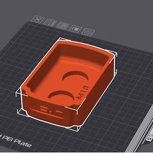 Pode incluir: Um molde de plástico vermelho com duas depressões circulares e o texto "M13" impresso na lateral. O molde está sobre uma superfície preta texturizada.
