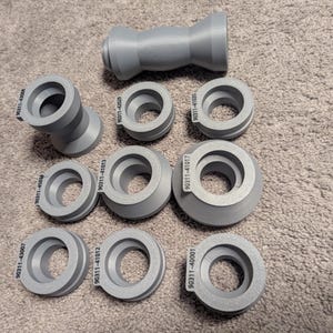 May include: A collection of gray, 3D-printed industrial parts, including cylindrical and angled components. Each part has a unique identification number printed on its surface. The parts are arranged on a textured, neutral-toned surface.