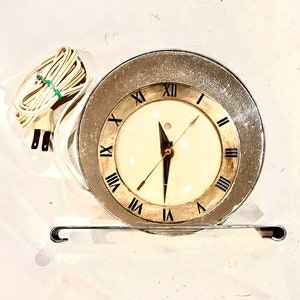 以下が含まれることがあります： ローマ数字の文字盤が付いた銀と金の時計。時計は白いコードで差し込み式で、金属製のスタンドが付いています。