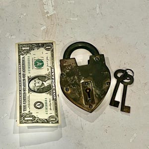 May include: An antique brass padlock with two keys and a one-dollar bill. The padlock is heart-shaped with a curved handle. The keys are small and metallic. The dollar bill is folded and shows the portrait of George Washington.