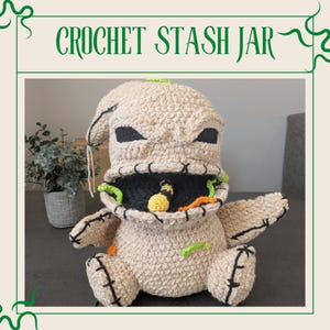 May include: A beige crochet stash jar shaped like a character with black eyes and mouth, and green and orange accents. The jar has a wide open mouth and is sitting on a dark surface. The text "CROCHET STASH JAR" is at the top.