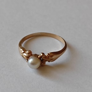 May include: A gold-colored ring with a central, round, white pearl. The ring features decorative, leaf-like accents on either side of the pearl. The band is thin and smooth, creating a classic and elegant design.