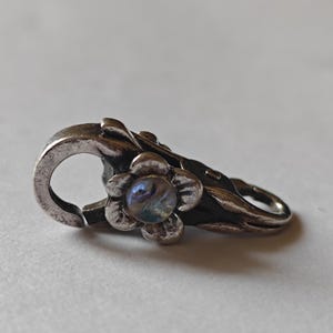 Peut inclure: Un fermoir argenté avec un motif floral. Le fermoir présente une fleur avec un centre nacré et des accents de feuilles détaillés. Le fermoir est conçu pour sécuriser des bijoux ou d'autres accessoires.