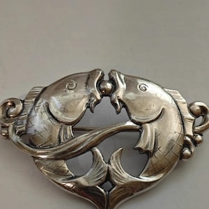 May include: A silver brooch shaped like two fish intertwined, with their tails forming a heart shape.