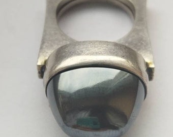 May include: A silver ring with a dark gray, oval-shaped gemstone. The ring has a square band with a raised edge.