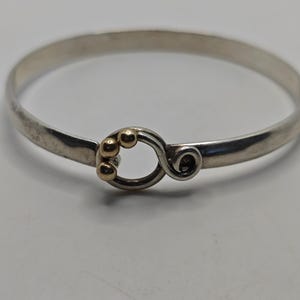 May include: A silver bangle bracelet with a gold accent in the shape of a swirl with three gold beads.