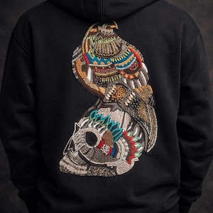 Puede incluir: Sudadera con capucha negra con un diseño bordado de un águila posada sobre una calavera. El águila y la calavera están adornadas con abalorios coloridos en tonos marrones, azules, rojos y verdes. La sudadera es de tejido suave y oscuro.