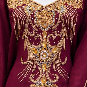 May include: A burgundy-colored garment with intricate gold embroidery and embellishments. The V-neckline and central panel feature elaborate floral and geometric patterns, with dangling bead accents. The design incorporates golden beads and gemstones.