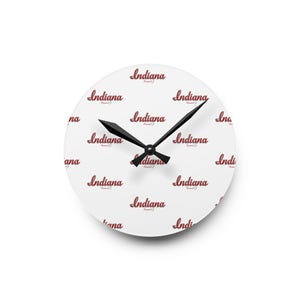 May include: A round white wall clock with black hands. The clock face features the word "Indiana" in a repeating pattern in a red script font. The clock hands are black.