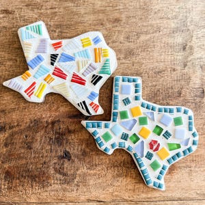 Puede incluir: Dos posavasos de mosaico con forma de Texas. Uno presenta un diseño de mosaico colorido con azulejos rojos, amarillos, azules y verdes. El otro tiene un diseño similar con una paleta de colores diferente.
