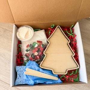 May include: A craft kit in a cardboard box, featuring a wooden Christmas tree cutout, mosaic tiles, glue, a sponge, gloves, and stir sticks. The box is filled with red and green paper shreds.