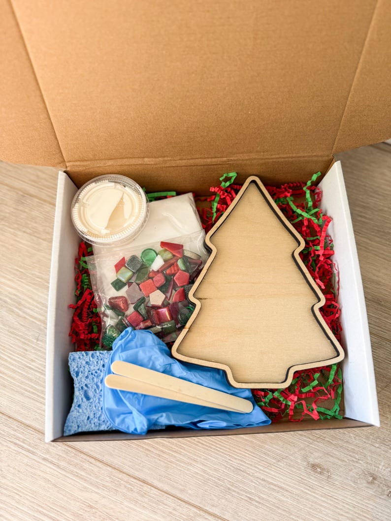 May include: A craft kit in a cardboard box, featuring a wooden Christmas tree cutout, mosaic tiles, glue, a sponge, gloves, and stir sticks. The box is filled with red and green paper shreds.