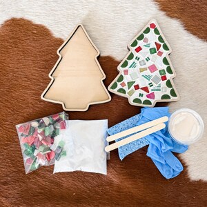 May include: A Christmas tree mosaic kit with two wooden tree-shaped bases, one decorated with colorful mosaic tiles. The kit includes bags of tiles, grout, a sponge, wooden sticks, and gloves. The items are arranged on a brown and white patterned surface.