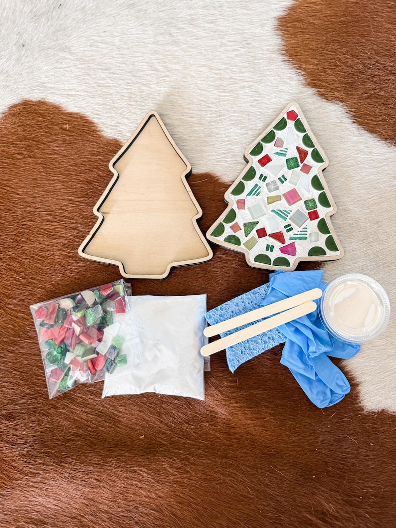 May include: A Christmas tree mosaic kit with two wooden tree-shaped bases, one decorated with colorful mosaic tiles. The kit includes bags of tiles, grout, a sponge, wooden sticks, and gloves. The items are arranged on a brown and white patterned surface.