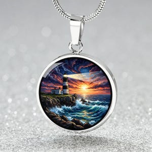 May include: A silver-toned pendant necklace featuring a round charm with a lighthouse scene. The charm depicts a lighthouse on a rocky coast with a sunset sky, ocean waves, and a bright beam of light. The necklace has a snake chain.